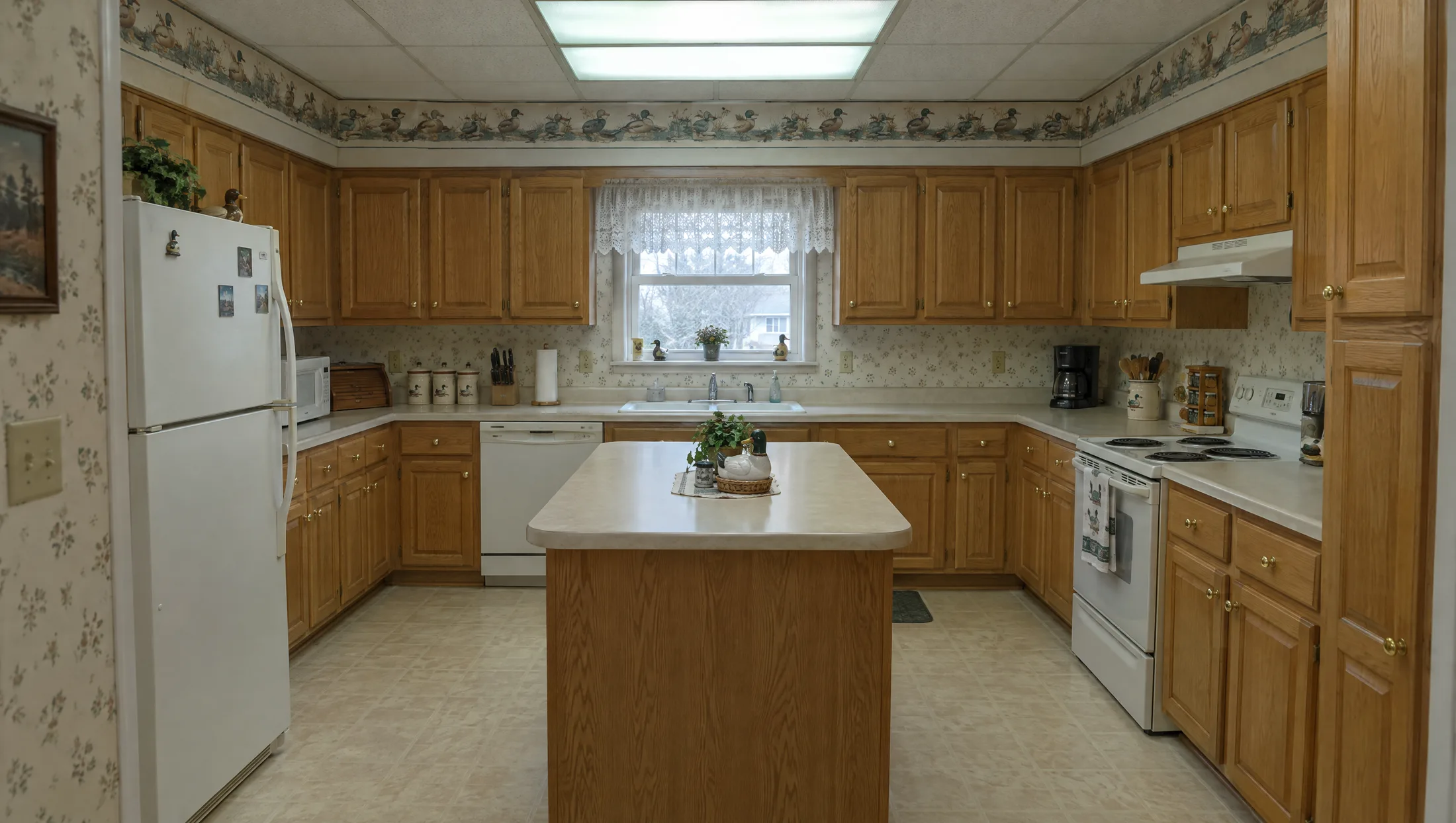 An outdated 1994 kitchen with honey oak cabinets and duck wallpaper, transforming into a white oak and Calacatta marble kitchen